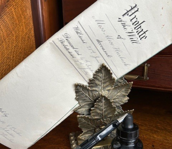 document titled Probate of the Will resting on a wooden surface next to a decorative leaf-shaped holder and an ink bottle featuring a pen symbolizing legal matters and historical records
